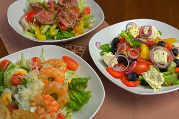 Close-up of three gourmet salads: shrimp Caesar, Greek salad with feta and olives, and beef with sesame on fresh vegetables, served on white plates with a rustic table setting.