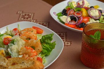 Close-up of shrimp Caesar salad with cherry tomatoes and Parmesan, next to a Greek salad and a glass of iced tea with mint on embroidered table linen.