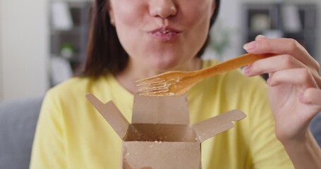 Happy hungry woman eating takeout Italian pasta, holding wooden fork and eco paper box to eat gourmet lunch. Girl with plastic free sustainable utensils enjoying spaghetti with creamy sauce at home