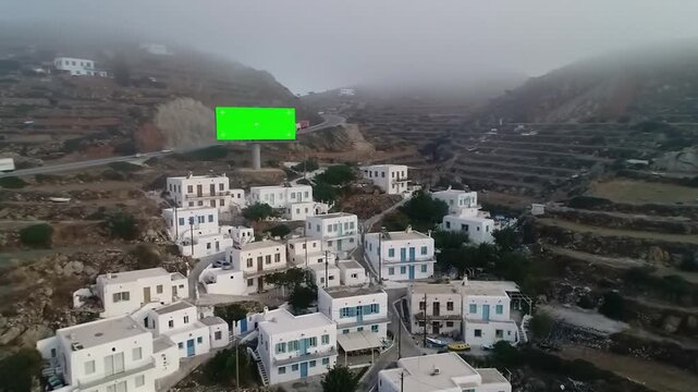 Aerial view of a traditional Greek village with a large green screen billboard on a misty hillside.