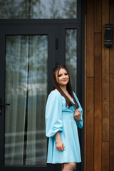 A woman stands on a veranda near a wooden wall of a building. She is wearing a blue dress with white polka dots and white sneakers