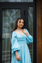 A woman stands on a veranda near a wooden wall of a building. She is wearing a blue dress with white polka dots and white sneakers