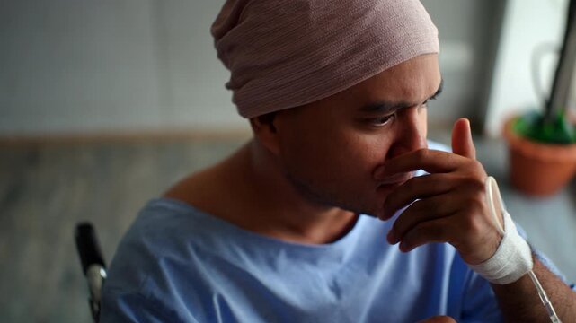 Depressed young patient man with cancer in headscarf feeling stressed and hopeless while sitting alone on wheelchair in the hospital room, mental health and disease awareness on world cancer day.
