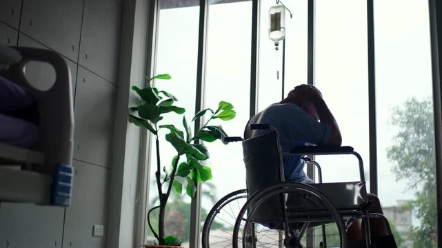 Rear view of depressed patient man with cancer sitting on wheelchair suffering headache and looking out the window at hospital, sickness male wearing head scarf feeling unwell and lonely.