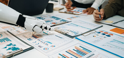 A modern business scene featuring a robotic hand assisting a professional team in analyzing charts, graphs, and reports on a collaborative workspace.Noogenesis