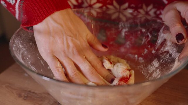 Close Up of Woman Kneading Dough with Red Coloring in Bowl While Wearing Holiday Sweater in Warm Kitchen Lighting