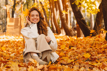 Smiling young woman sits in the autumn foliage on the ground of the forest with the yellow leaves