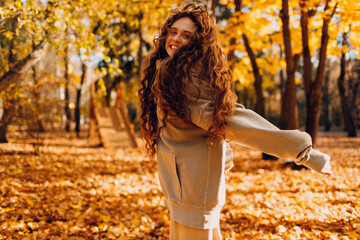 Smiling young woman enjoys the autumn weather in the fall park with the yellow leaves at sunset