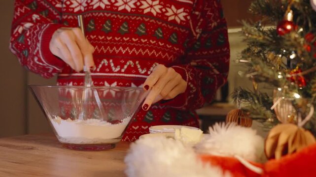 Woman Stirring Flour in Glass Bowl with Whisk on Kitchen Table Decorated with Christmas Tree and Ornaments