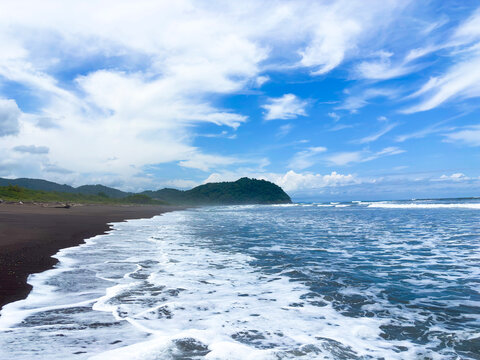 Wide view of Camaronal Beach in Costa Rica, with waves breaking on dark sand under a blue sky filled with clouds.