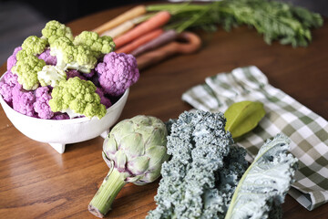 Fresh organic vegetables on a wooden table. Purple cauliflower, Romanesco broccoli, kale, and artichoke with rainbow carrots in the background.