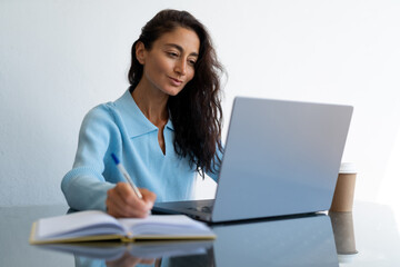 Thoughtful middle-aged woman with long wavy dark hair in a light blue sweater writing in a notebook while working on a laptop at a glass desk with takeaway coffee in a bright minimalist office