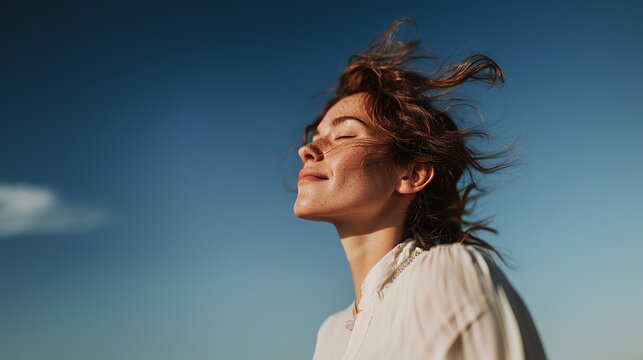 A woman with closed eyes enjoys the fresh air, hair blowing in the breeze under a clear blue sky.