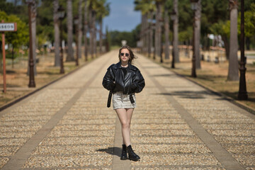 Fototapeta premium Young, beautiful, blonde woman, with leather jacket, snake miniskirt, boots and sunglasses, posing with her hands in her pockets, solitary and independent, in the middle of an avenue.
