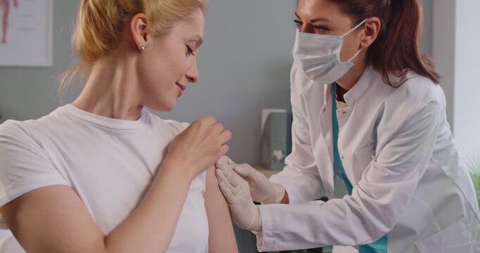 Female doctor in protective mask giving injection to adult woman patient in clinic. Friendly nurse administering vaccine into upper arm. Immunization, preventive treatment and health care concept.