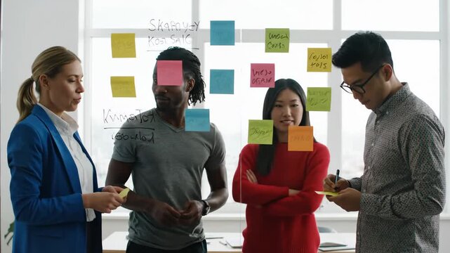Diverse Team Brainstorming on Glass Wall - A diverse group of four professionals collaborates on a project, using sticky notes and writing on a transparent glass wall in a modern office.