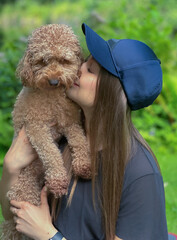 Young woman with long hair wearing cap kisses her poodle dog outdoors