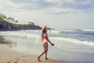 Confident woman with braids strolls barefoot along shore, holding smartphone as symbol of freedom, digital self-expression and personal empowerment in movement.