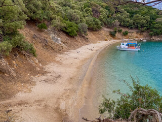 a small white fishing boat floating in a secluded bay with clear blue-green water and a sandy beach, surrounded by a steep, wooded bank, travel and tourism.