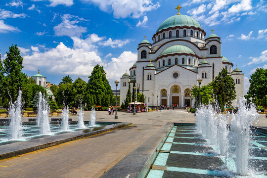 The Church of Saint Sava in Belgrade, a stunning example of Byzantine architecture, shines under a blue sky with white clouds. Greenery and fountains surround the square. Serbia