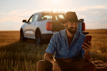 Happy farm worker using cell phone while relaxing in field at sunset.