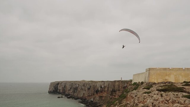 Paraglider flying above Fortaleza de Sagres and rugged cliffs, Algarve, Portugal