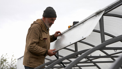 An adult man installs an arched greenhouse. He attaches cellular polycarbonate to a metal frame.