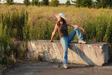 Fototapeta premium Beautiful young woman in jeans and hat sitting on a concrete block by the road