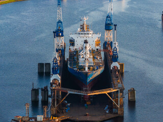 A large ship in a dry dock surrounded by two blue and white cranes, with scaffolding and equipment indicating maintenance work in Riga, Latvia.