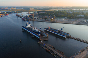 Naklejka premium A shipyard in Riga, Latvia, features large vessels in dry docks, cranes, and industrial structures. Sunset lighting highlights greenery and urban areas.