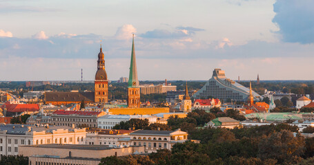 Obraz premium Riga Cathedral and St. Peter's Church spires rise above the skyline, with the National Library of Latvia in the background under a golden sunset glow.