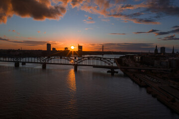 Fototapeta premium The sun sets over Riga, Latvia, with the Railway Bridge spanning the Daugava River. The Riga TV Tower and old town skyline are visible in the distance.