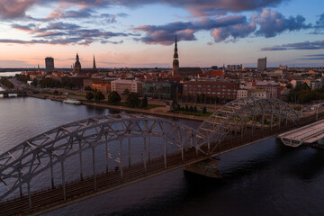 Riga, Latvia at sunset with the Daugava River, Railway Bridge, and Old Town landmarks like St. Peter's Church and Riga Cathedral in the background.