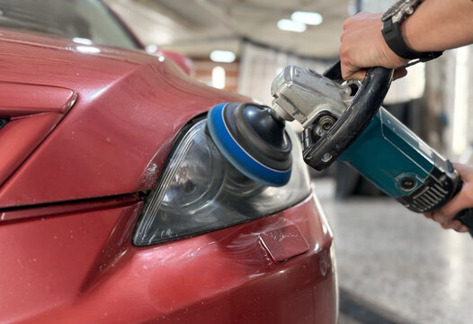 Car detailing series: The process of polishing the headlights. Hands of male professional worker with orbital polisher in auto repair shop, polishing car headlight. Car wax polishing process close up.