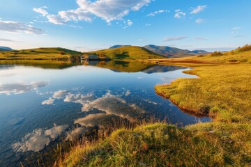 Obraz premium Serene lake mirroring the sky with grassy shores and distant hills under a bright cloudy sky view landscape