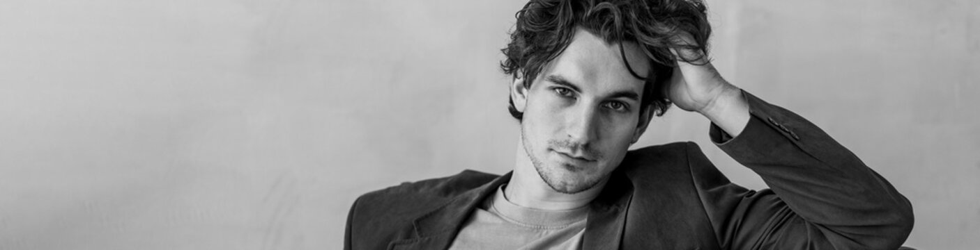 Banner. Young sexy handsome fit man in a brown suit looking at the camera sitting on a chair in the studio. Grey background, loft interior, studio. Causal stylish men's clothes, watch