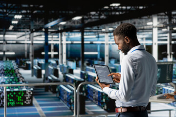 African american admin analyzing infrastructure performance outputs on tablet while he stands on industrial platform. Server room employee finetuning mainframes configurations.