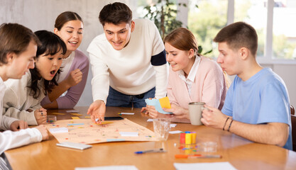 Cheerful adult students playing board game excitedly sitting around the table