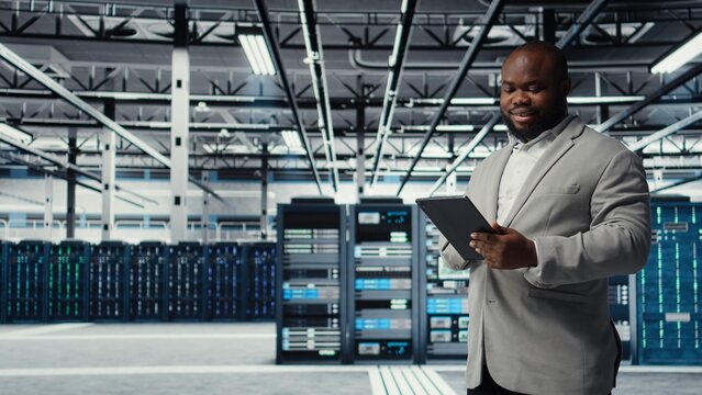 Cheerful data center programmer looking at code debugging tools running on tablet. Server farm IT professional typing on device touchscreen, writing script automating tasks, camera A