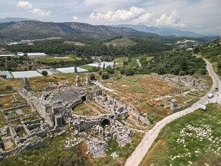Tlos theatre and agora lie beside a winding rural road in Mugla, Turkey with forested hills and cumulus clouds Visitors explore stepped seating and vaulted passages cut into the slope above the valley