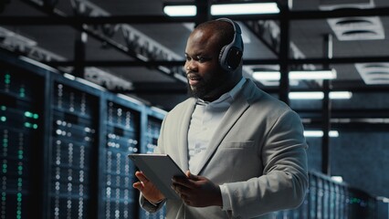 Server room programmer wearing headphones troubleshooting errors using software on tablet. IT specialist listening music, walking in data center fixing bugs using device , camera B
