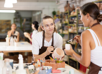 Young man sits near table and decorates clay ceramic craft vase, plate, works in company of like-minded people. Hand made art, hobbies and creativity.