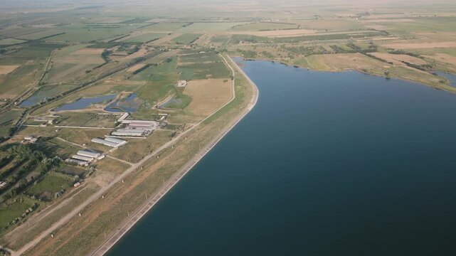 Dam of Djizak Reservoir is visible near agricultural buildings and ponds. Water is calm, bordered by fields. Roads and farmland blend with nature, showing connection between infrastructure and land