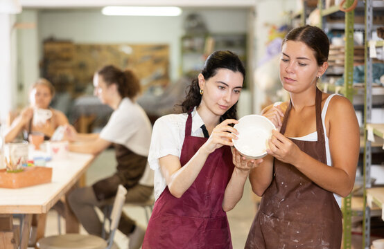 Girl holds ceramic bowl in hands and shows it to friend. Two visitors of courses art workshop, look at painted product, admire result of work. Girls inspect painted ceramic product