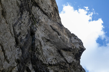 A dramatic low-angle view of a towering limestone cliff face in Romania's Bicaz Canyon (Cheile Bicazului), with intricate rock textures set against a bright blue sky with white clouds.