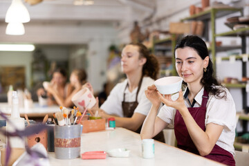 Interested young woman in pinafore drawing on handmade clay jar with paintbrush in art studio