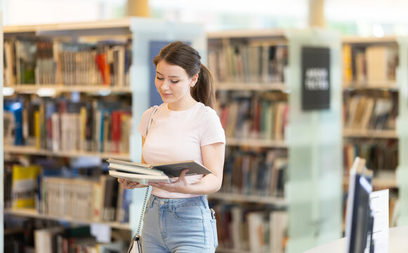 Young female student reading book standing in library