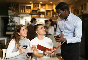 In dim light of restaurant hall, African male waiter communicates with friends customers, answers questions about composition of dish of day. Couple in restaurant makes order to waiter