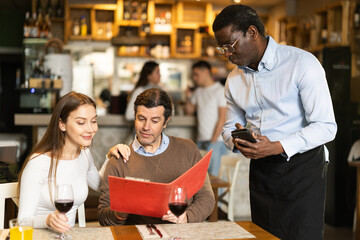 African waiter tells a European couple about the choice of dishes and the range of wines in the restaurant. Joyful couple came on a date and make an order from the waiter
