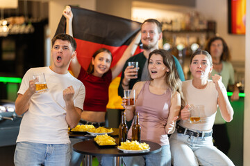 Group of fans of Germany team with a flag are watching the match on TV in a beer bar. They are worried and supporting their